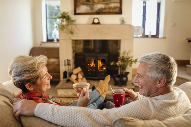 How to Keep Warm During Extreme Cold Weather. Mature couple sat infront of a fire drinking hot chocalate from mugs.