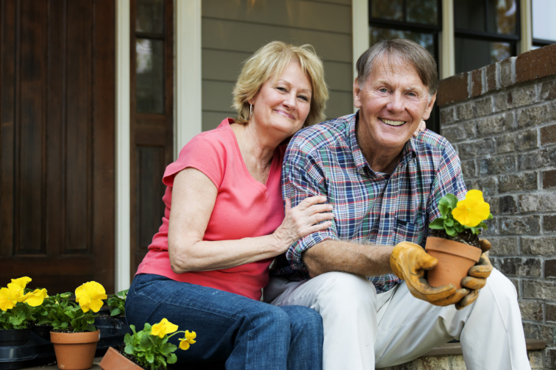 Why Buying a Heat Pump Instead of a Furnace Makes Sense. Senior Man And Woman Sit Outside Their House And Smile At The Camera Holding Pot Plant.